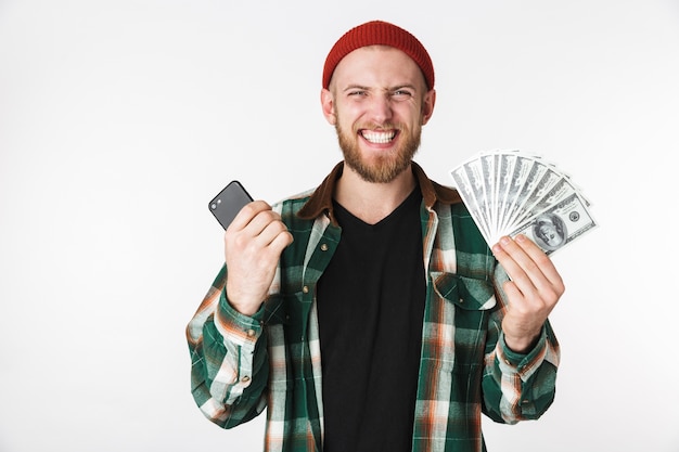 Imagen de chico positivo vistiendo camisa a cuadros sosteniendo teléfono celular y dinero en dólares, mientras está de pie aislado sobre fondo blanco.