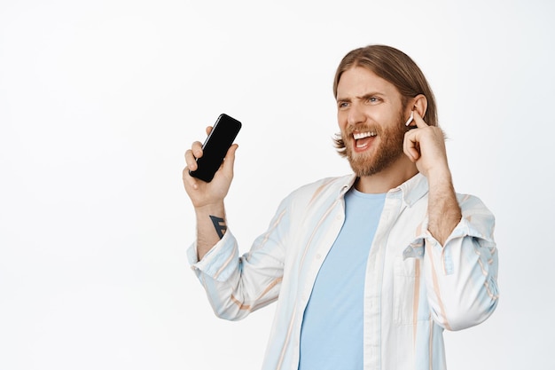 Imagen de un chico rubio feliz bailando con auriculares, escuchando música en auriculares, sosteniendo un teléfono móvil y sonriendo complacido, de pie sobre un fondo blanco.