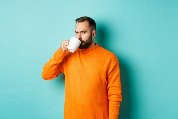 Imagen de un chico tomando café y mirando sospechoso a la cámara, mirando algo extraño, de pie con un suéter naranja sobre fondo turquesa