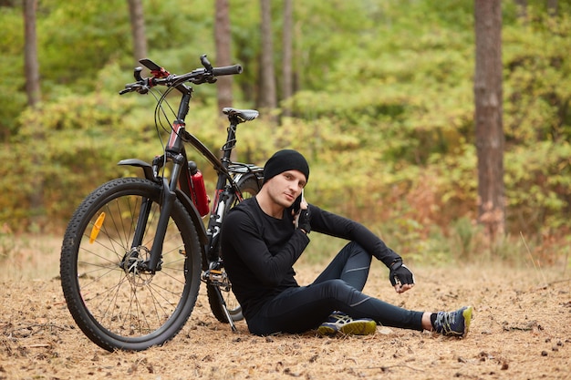 Imagen de un ciclista de montaña masculino descansando en un viaje en bicicleta, sentado en el suelo bajo un árbol con su bicicleta acostada junto a él