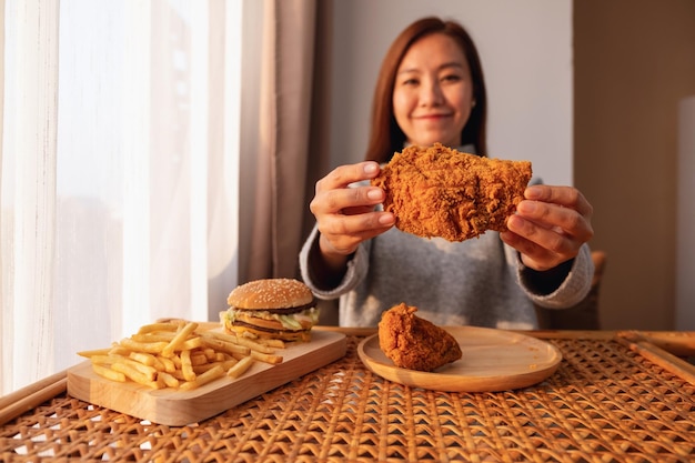 Imagen de cierre de una joven asiática sosteniendo y comiendo pollo frito con hamburguesa y papas fritas en la mesa en casa