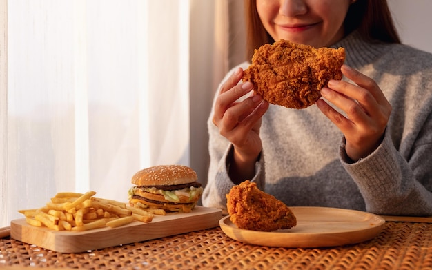 Imagen de cierre de una joven asiática sosteniendo y comiendo pollo frito con hamburguesa y papas fritas en la mesa en casa