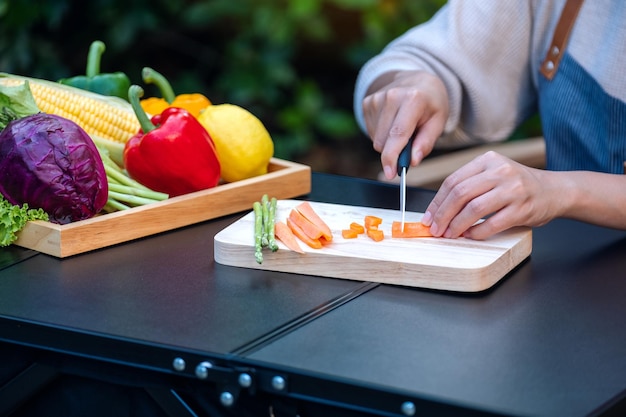 Imagen de cierre de una mujer cortando y picando zanahoria con un cuchillo en una tabla de madera con verduras mixtas en una bandeja