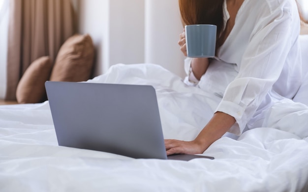 Imagen de cierre de una mujer que trabaja en una computadora portátil y toma café mientras se sienta en una cama en casa