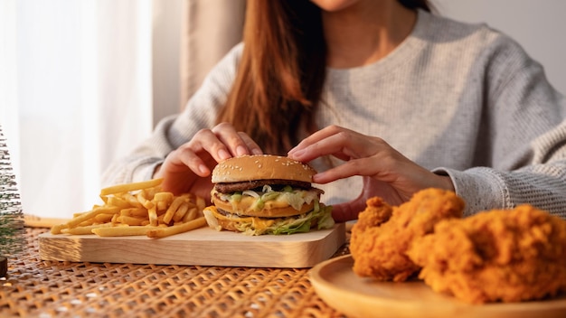 Imagen de cierre de una mujer sosteniendo y comiendo hamburguesas y papas fritas con pollo frito en la mesa en casa