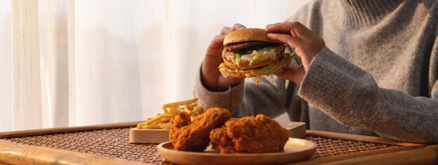 Imagen de cierre de una mujer sosteniendo y comiendo hamburguesas y papas fritas con pollo frito en la mesa en casa