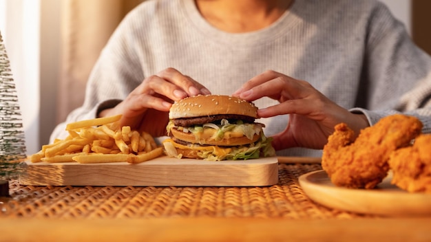 Imagen de cierre de una mujer sosteniendo y comiendo hamburguesas y papas fritas con pollo frito en la mesa en casa