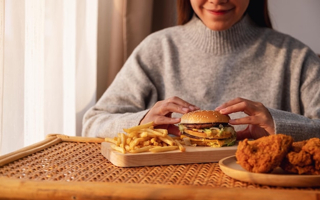 Imagen de cierre de una mujer sosteniendo y comiendo hamburguesas y papas fritas con pollo frito en la mesa en casa