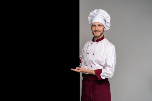 Imagen del cocinero joven sonriente en uniforme que se encuentran aisladas sobre fondo gris de la pared. Mirando la cámara apuntando.