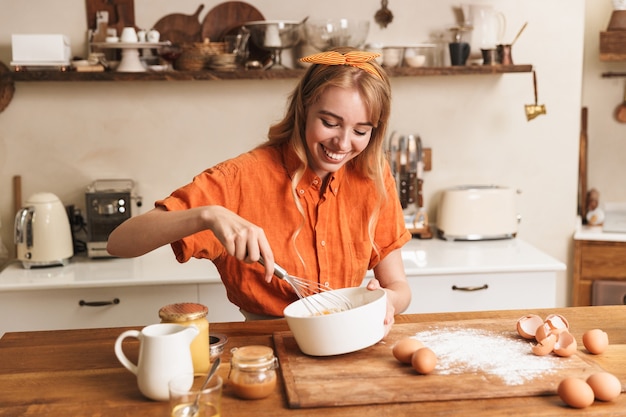 Imagen de un cocinero de la muchacha rubia joven sonriente alegre feliz que cocina en la cocina.