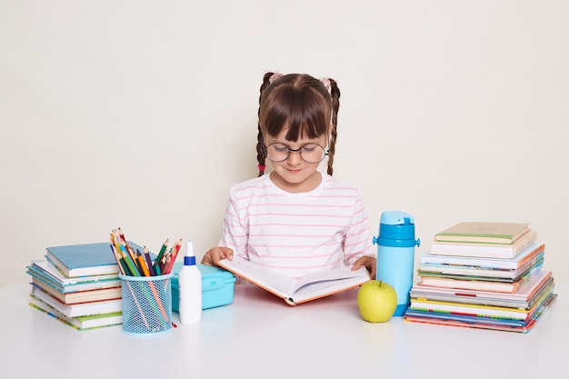 Imagen de una colegiala seria y atenta con cabello oscuro y trenzas sentada en la mesa rodeada de libros y leyendo haciendo tareas de literatura durante la lección