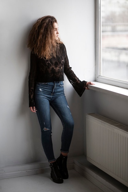 Foto imagen completa de una hermosa joven con el pelo rizado, blusa negra y jeans posando cerca de las ventanas