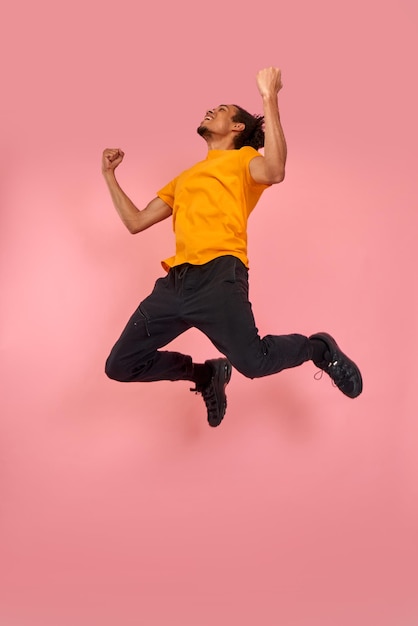 Imagen completa de un hombre negro divertido y alegre con zapatillas de deporte, loco volando sin preocupaciones saltando con los brazos levantados, celebrando la victoria, divirtiéndose, descansando, relajándose y disfrutando, aislado en un fondo rosa