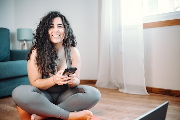 Imagen completa de una joven mujer con curvas en ropa deportiva sentada en el suelo descansando mientras hace yoga en casa. usando el chat telefónico. Bajar de peso en casa. Clase en línea con el concepto de computadora portátil.
