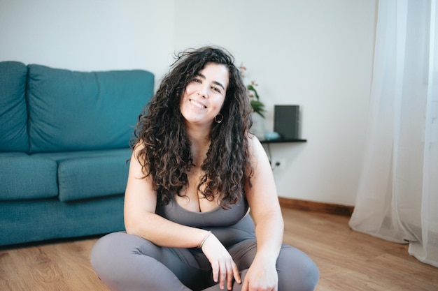 Imagen completa de una joven mujer latina con curvas en ropa deportiva sentada en el suelo descansando mientras hace yoga en casa, mujer latina. Felicidad nuevo hábito saludable. Clase en línea con el concepto de computadora portátil.