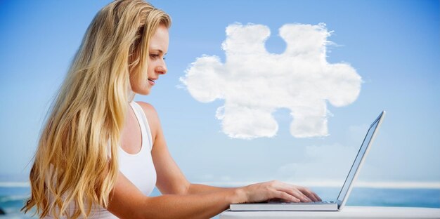 Imagen compuesta de una guapa rubia usando su laptop en la playa contra la pieza del rompecabezas en la nube