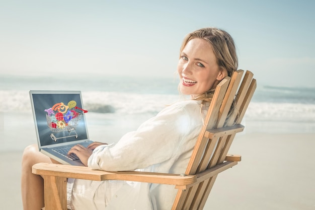 Imagen compuesta de una hermosa rubia sentada en una tumbona usando una laptop en la playa