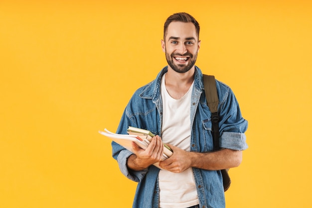 Foto imagen de contenido estudiante chico en ropa de mezclilla sonriendo mientras sostiene libros de ejercicios aislado sobre pared amarilla