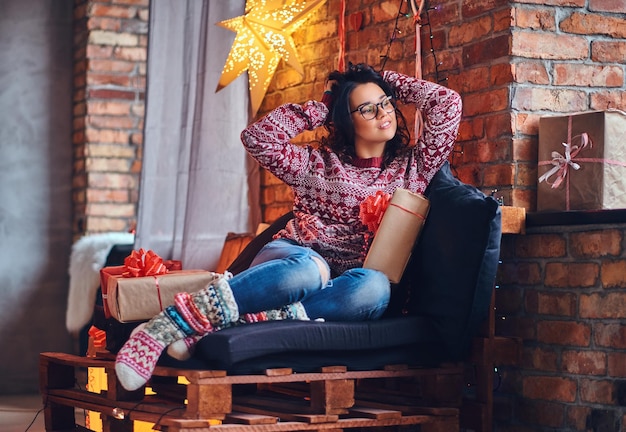 Imagen corporal completa de una mujer morena con anteojos vestida con jeans y un suéter rojo posando en un sofá de madera en una habitación con decoración navideña.