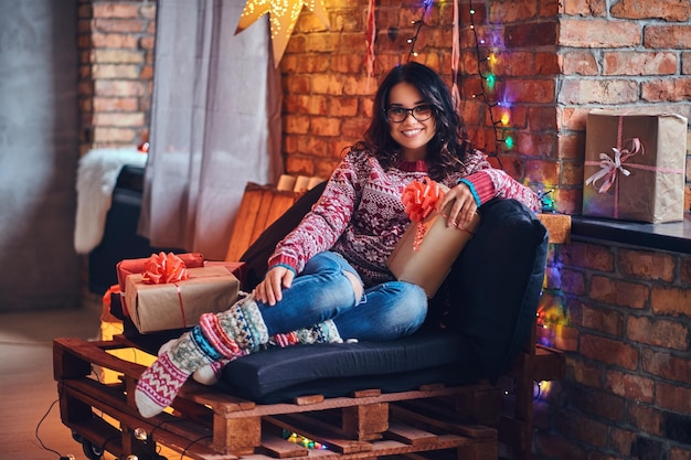 Imagen corporal completa de una mujer morena con anteojos vestida con jeans y un suéter rojo posando en un sofá de madera en una habitación con decoración navideña.