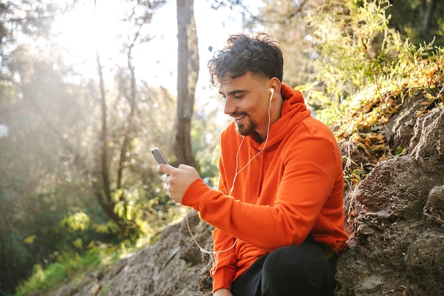 Imagen de corredor de hombre guapo feliz joven deportes fitness al aire libre en el parque escuchando música con auriculares mediante teléfono móvil.