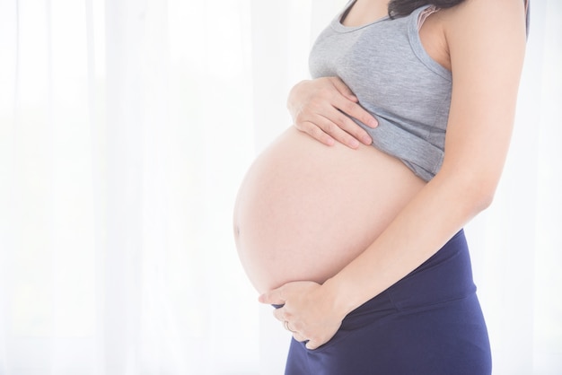 Foto imagen cosechada de la mujer embarazada asiática, sosteniendo su vientre delante de la ventana brillante.