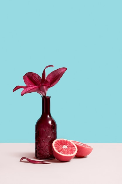 La imagen creativa de la naturaleza muerta con el florero de la botella, la flor roja y el pomelo en la tabla gris se colocan en fondo azul. Imagen conceptual para tienda de flores con espacio de copia para el diseño. tarjeta de felicitación.