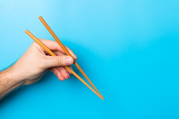 Imagen creativa de palillos de madera en manos masculinas sobre fondo azul. Comida japonesa y china con espacio de copia