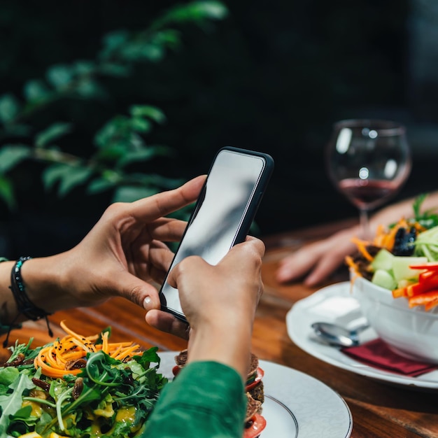 Imagen cuadrada de una mujer joven fotografiando una deliciosa hamburguesa vegetariana y una ensalada orgánica mixta en un plato blanco en un restaurante vegetariano