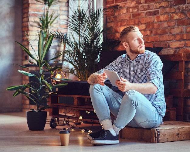 La imagen de cuerpo completo de un hombre rubio y elegante vestido con una camisa de lana y jeans usando una tableta en una sala de estar.