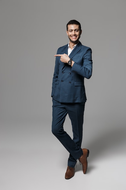 Imagen de cuerpo entero del atractivo hombre de negocios árabe de 30 años en traje formal posando con el dedo a un lado, aislado sobre la pared gris