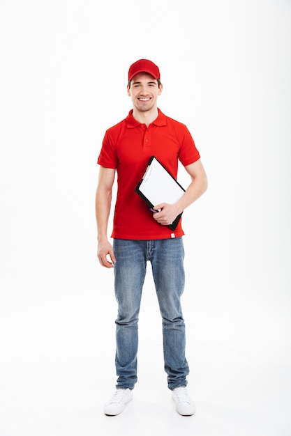 Imagen de cuerpo entero del joven mensajero 20s en uniforme rojo y jeans mirando a la cámara y sosteniendo el portapapeles, aislado sobre un espacio en blanco