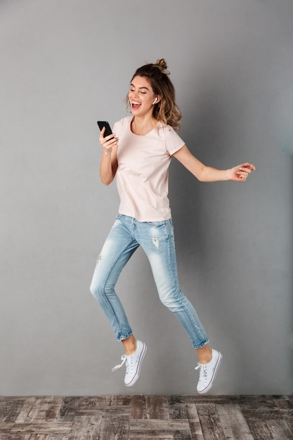 Imagen de cuerpo entero de mujer alegre en camiseta escuchando música desde un teléfono inteligente con auriculares mientras salta y se divierte en gris