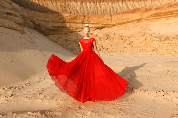 Imagen de cuerpo entero de una mujer joven en vestido rojo que agita con tela voladora en el fondo de arenas doradas.