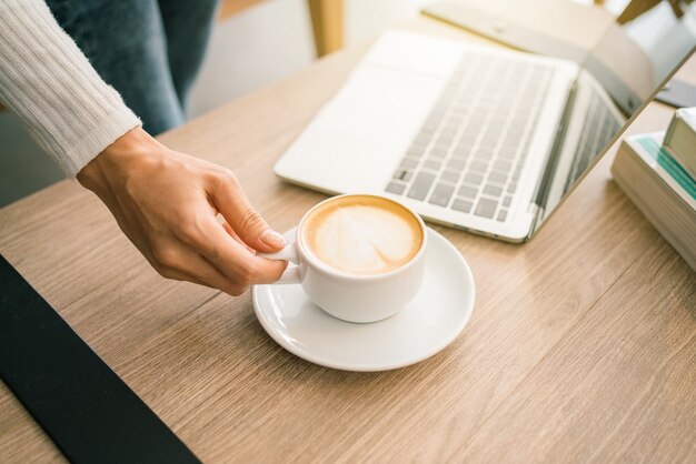 Imagen de cultivo de una mano de mujer con un suéter sosteniendo una taza de café con leche caliente por la mañana, portátil.