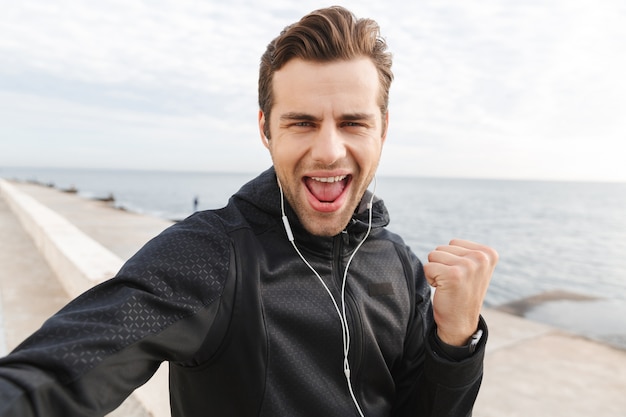 Imagen de deportista alegre de 30 años en ropa deportiva negra y auriculares, tomando una foto selfie en el teléfono móvil mientras camina a la orilla del mar