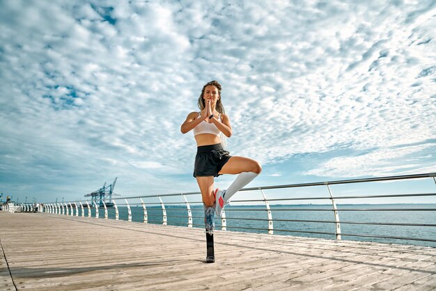 Imagen de una deportista discapacitada joven y fuerte que hace ejercicio de yoga en un puente cerca del mar