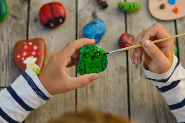Una imagen detallada de las manos de un niño dibujando un búho verde en una piedra con pinturas acrílicas Los pasatiempos domésticos son auténticos Obras de arte en piedras