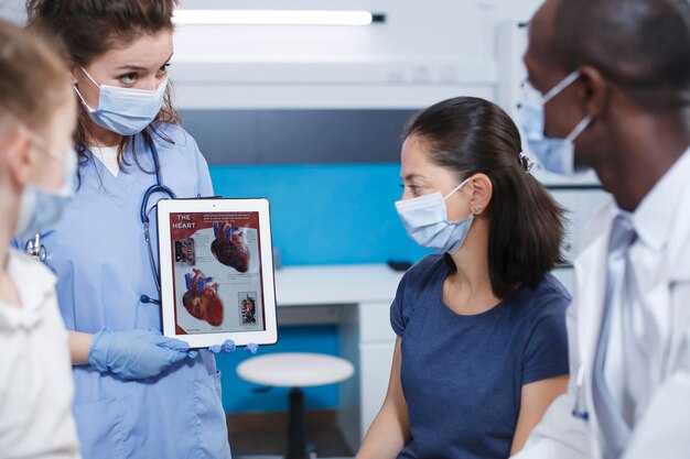 Imagen detallada de profesionales de la salud usando una tableta para explicar a las pacientes femeninas sobre el corazón humano La enfermera sostiene un dispositivo mientras el médico masculino describe la importancia de los exámenes médicos