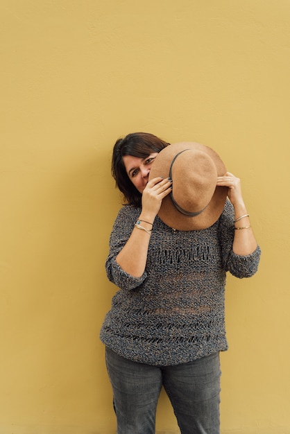 Una imagen divertida de una mujer mirando a la cámara mientras sostiene un sombrero en sus manos y se apoya contra una pared de color amarillo mostaza. Copia espacio