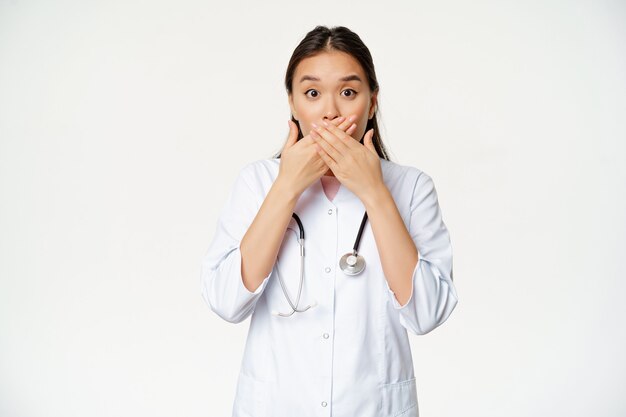 Imagen de la doctora asiática sorprendida cubre sus labios con las manos, cierra la boca y parece preocupado, gesto tabú, de pie sobre fondo blanco.