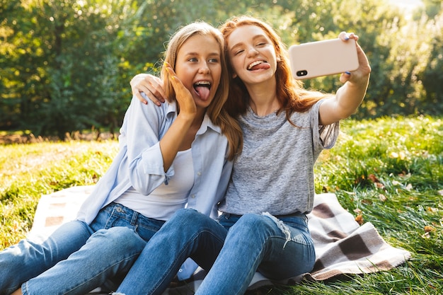 Imagen de dos amigas felices vistiendo jeans sentados en el césped y tomando selfie en smartphone, mientras descansa en el parque de verano