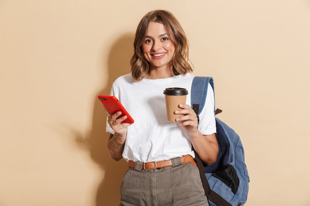 Imagen de una elegante chica estudiante sosteniendo una taza de café para llevar y un teléfono inteligente aislado sobre una pared beige