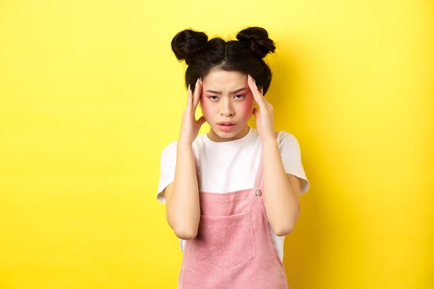 Imagen de una elegante mujer japonesa con maquillaje brillante, que tiene dolor de cabeza, toca la cabeza y parece cansada, tiene migraña dolorosa, amarilla.