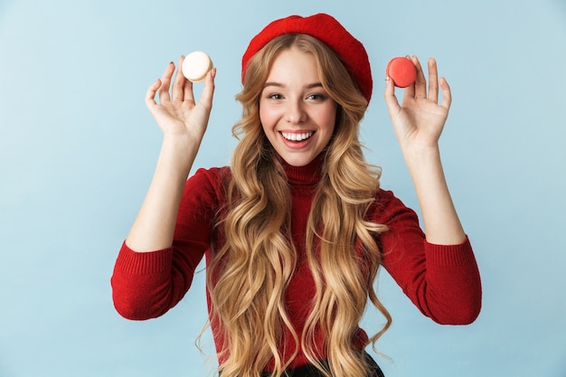 Imagen de la elegante mujer rubia de 20 años vistiendo boina roja sosteniendo galletas macaron aislado