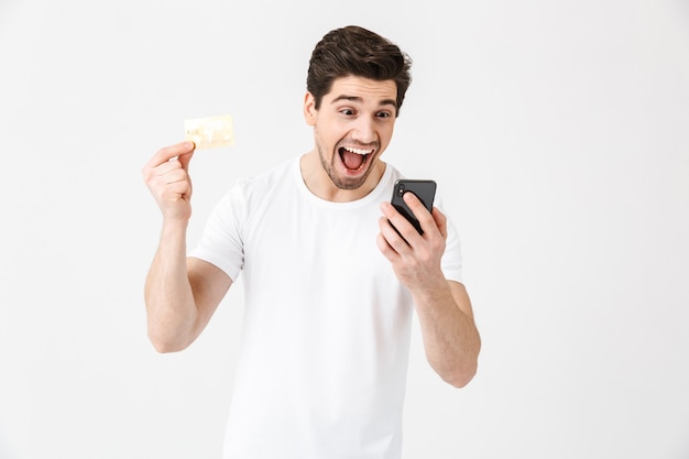 Imagen de emocionado emocional joven feliz gritando posando aislada sobre pared blanca con tarjeta de crédito de explotación de teléfono móvil.