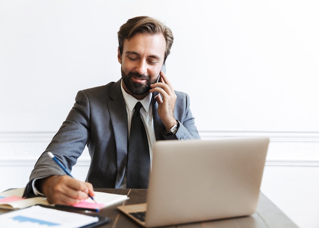 Imagen del empresario sin afeitar sonriente vistiendo traje formal hablando por teléfono celular y anotando notas mientras trabaja en la computadora portátil en la oficina