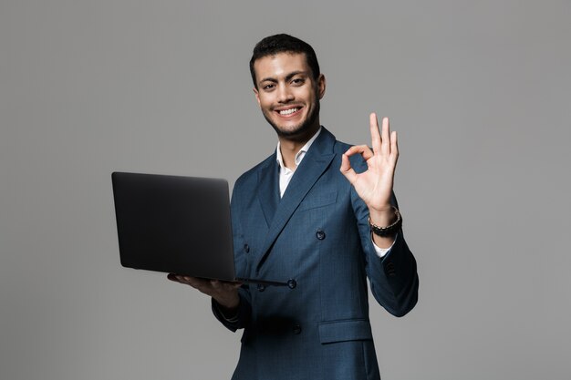 Imagen del empresario árabe positivo de 30 años en traje formal sonriendo y sosteniendo un portátil, aislado sobre una pared gris