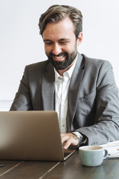 Imagen del empresario confiado riendo vistiendo traje formal escribiendo en la computadora portátil y tomando café mientras trabaja en la oficina