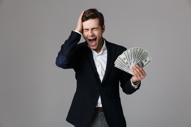 Imagen del empresario emocionado de 30 años en traje sonriendo y sosteniendo el ventilador de dinero en billetes de dólar, aislado sobre pared gris